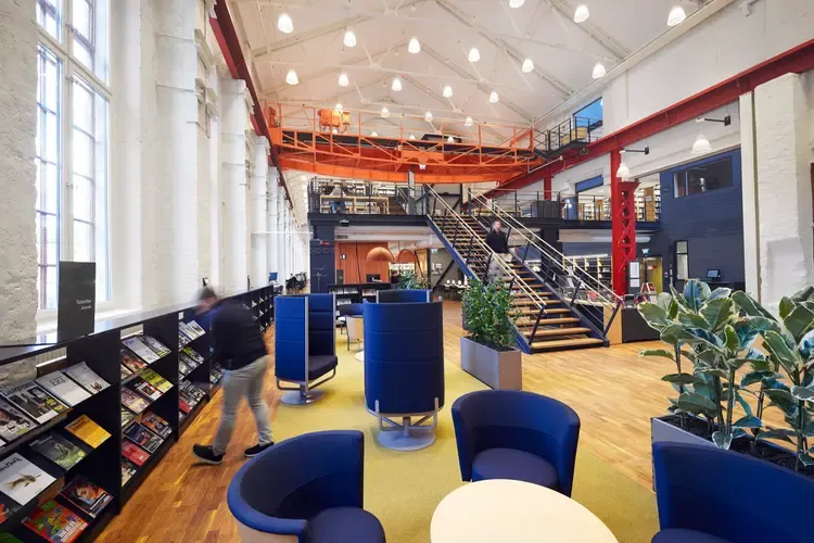 Bibliotekets tidskriftshörna med snurrfåtöljer, bord och gröna växter. Längs väggen bredvid syns tidskrifter på rad.